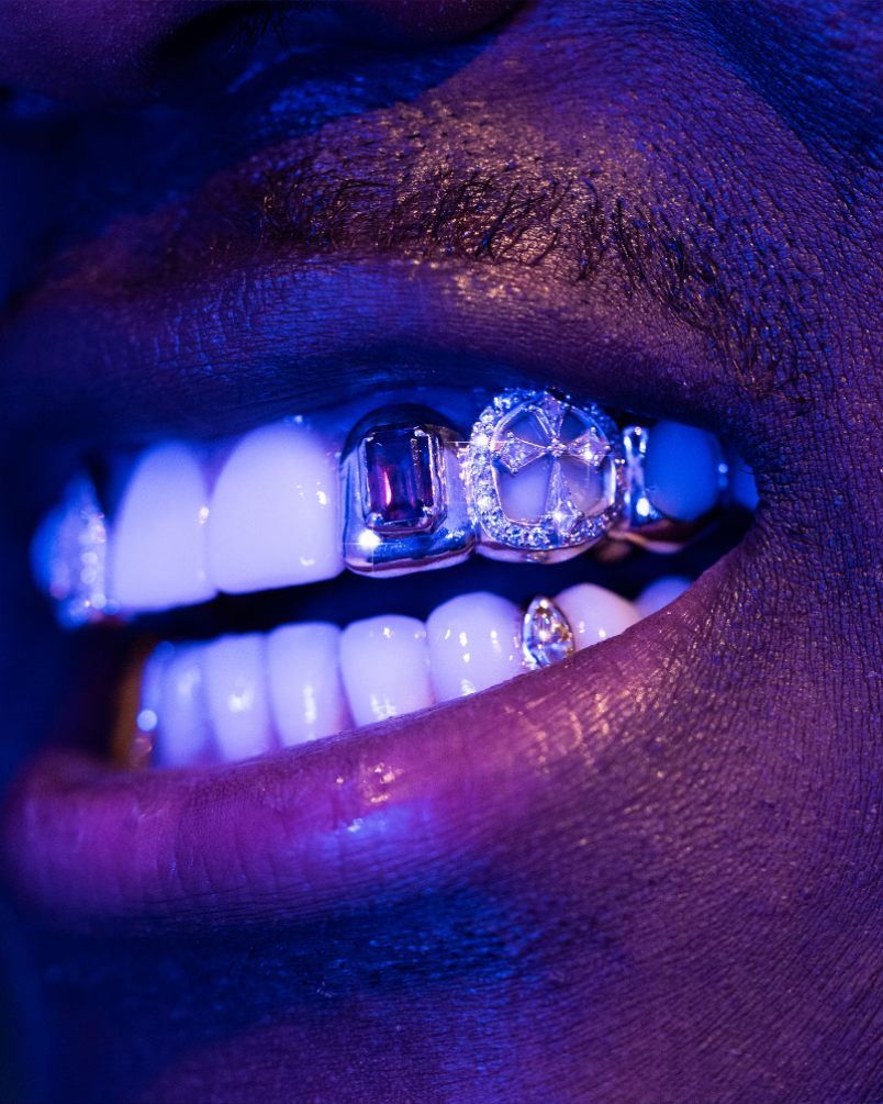 Close-up of a mouth with diamond-encrusted grillz under a purple light.